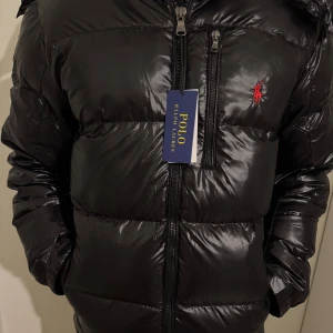 Svart pufferjacka från Polo Ralph Lauren (Glans) - Snygg svart pufferjacka från Polo Ralph Lauren med dragkedja och ikonisk logga på bröstet. Jackan har en glansig finish och är perfekt för kyliga dagar. Den har långa ärmar och en bekväm passform. [ÄKTHETSBEVIS FINNS]