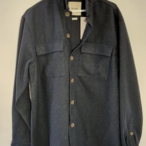 Blå overshirt från Only & Sons - Snygg blå overshirt från Only & Sons i loose fit. Tillverkad av 75% polyester och 25% akryl, perfekt för en stilren look. Jackan har knappar framtill och två bröstfickor för extra stil och funktionalitet. Ursprungligt pris var  900 SEK. 