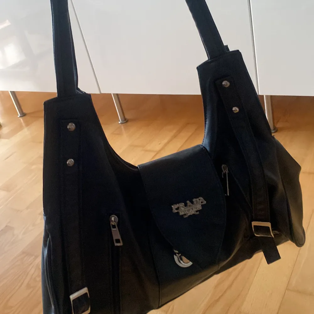 Snygg svart axelväska i skinn med flera dragkedjor och ett spänne framtill. Väskan har dubbla handtag och ett rymligt fack med mönstrat foder. Perfekt för att bära med sig alla nödvändigheter.. Laukut & Käsilaukut.
