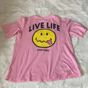 Rosa t-shirt från SmileyWorld x H&M - Säljer en rosa t-shirt från SmileyWorld x H&M med ett stort gult smileytryck och texten 'LIVE LIFE' på baksidan. T-shirten är kortärmad och i mjukt bomullsmaterial. Perfekt för en avslappnad stil!