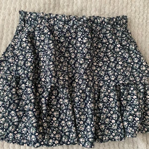 Blommig kjol från Basic Needs - Charmig blommig kjol från Basic Needs i mörkblått med små vita och rosa blommor. Kjolen har en volangdetalj och elastisk midja för bekväm passform. Perfekt för en somrig look!