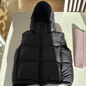 Svart dunväst från Zara - Snygg svart dunväst från Zara med huva och dragkedja. Perfekt för kyligare dagar med sin quiltade design och praktiska fickor. Västen är lätt och andas bra, vilket gör den idealisk för både höst och vinter.