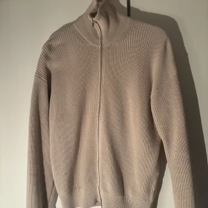 Beige kofta från Massimo Dutti - Säljer en stilren beige kofta från Massimo Dutti i 100% bomull. Koftan har en dragkedja framtill och en hög krage som ger en snygg look. Perfekt för lager-på-lager under kyligare dagar.