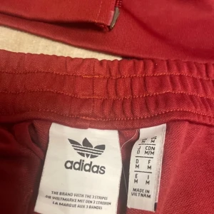 Röda träningsbyxor från Adidas - Snygga röda träningsbyxor från Adidas med de klassiska tre ränderna längs sidorna. Byxorna har en elastisk midja och är perfekta för sportiga aktiviteter. Matchande överdel ingår.