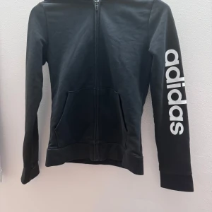 Svart hoodie från Adidas - Säljer en svart hoodie från Adidas med dragkedja och stor logga på ärmen. Tröjan har långa ärmar och en praktisk huva. Perfekt för en sportig look eller avslappnade dagar.