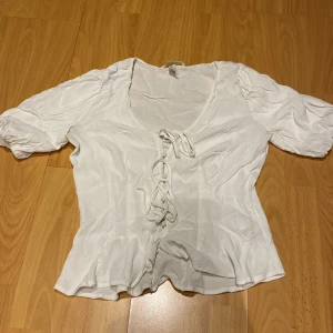 Vit blus från H&M - Säljer en söt vit blus från H&M med knytning framtill och korta puffärmar. Perfekt för en somrig look. Blusen har en v-ringad design och är tillverkad i ett lätt material.