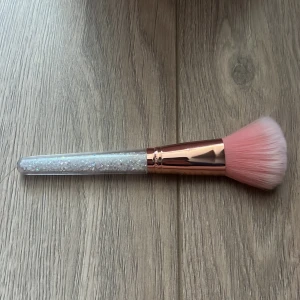 Glittrig sminkborste med rosa borst - En elegant sminkborste med ett glittrigt handtag och mjuka rosa borst. Perfekt för att applicera puder eller rouge med precision och stil. Handtaget har en skimrande effekt som ger en lyxig känsla.