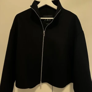 Svart jacka från Zara - Snygg svart jacka från Zara med dragkedja framtill. Perfekt för en stilren look. Jackan har långa ärmar och en hög krage som ger extra attityd.