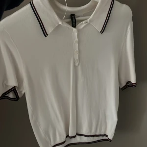 Vit pikétröja från Gina Tricot - Snygg vit pikétröja från Gina Tricot med svarta och röda detaljer på krage och ärmslut. Tröjan har korta ärmar och knappar framtill. Perfekt för en stilren look.