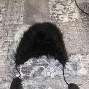 Svart pälsmössa - Säljer en svart pälsmössa med öronlappar och knytband. Perfekt för kalla vinterdagar när du vill hålla dig varm och stilren. Mössan har en fluffig och mjuk känsla.