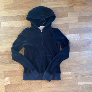 Svart hoodie från Juicy Couture - Snygg svart hoodie från Juicy Couture med texten 'JUICY LOVES NEW YORK' i guldpaljetter på ryggen. Tröjan har dragkedja och en mysig huva. Perfekt för en casual look med stil.