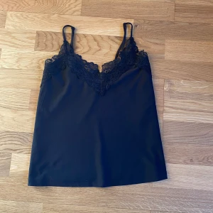 Svart linne med spetsdetaljer från Vero Moda - Snyggt svart linne från Vero Moda med vackra spetsdetaljer vid halsringningen. Perfekt för en stilren look. Linnet har smala axelband och är tillverkat i ett mjukt material.