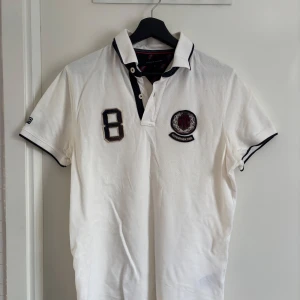 Vit pikétröja från Tommy Hilfiger - Snygg vit pikétröja från Tommy Hilfiger. Tröjan har korta ärmar och svarta detaljer vid krage och ärmslut. Perfekt för en sportig look! Originalpris 1000kr