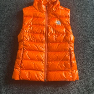 Orange dunväst från Moncler - Säljer en snygg samt unik orange dunväst från Moncler med dragkedja framtill. Västen har en hög krage och Moncler-logga på bröstet. Perfekt för att hålla värmen med stil! Nypris 11000kr