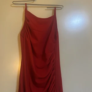 Röd långklänning med drapering - Elegant röd långklänning med smala axelband och vacker drapering framtill. Perfekt för en stilfull kväll. Klänningen har en smickrande passform och är tillverkad i ett mjukt material som faller fint.