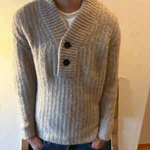 Beige stickad tröja från Burberry - Säljer en snygg beige stickad tröja från Burberry med två knappar framtill. Tröjan har en ribbad struktur och långa ärmar, perfekt för kyligare dagar. Den har en stilren design med en liten metallplatta med logotypen på framsidan.