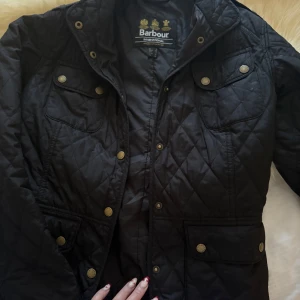 Svart quiltad jacka från Barbour - Säljer en stilren svart quiltad jacka från Barbour. Jackan har en klassisk design med knappar och flera praktiska fickor. Perfekt för höst och vår. Den har en normal passform och är långärmad.