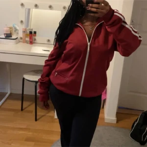 Röd träningsjacka från Adidas - Säljer en snygg röd träningsjacka från Adidas med vita detaljer. Jackan har dragkedja framtill och vita ränder längs ärmarna. Perfekt för en sportig look eller avslappnad stil. 🏃‍♂️