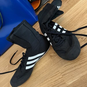 Svarta boxningsskor från Adidas - Säljer ett par svarta boxningsskor från Adidas med vita ränder. Skorna har hög snörning och är designade för optimal rörelse och stöd under träning.