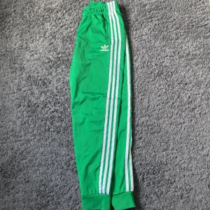 Adidas sst byxor - Snygga gröna träningsbyxor från Adidas med de klassiska tre vita ränderna längs sidorna. Byxorna har en elastisk midja och muddar vid bensluten för en bekväm passform. Perfekta för träning eller en avslappnad stil.