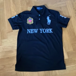 Svart pikétröja med New York-tryck - Snygg svart pikétröja från Ralph Lauren med ljusblått New York-tryck och nummer 5 på ryggen. Tröjan har korta ärmar och en klassisk krage. Den är dekorerad med ett broderat emblem och den ikoniska polospelaren på bröstet.
