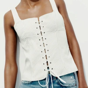 Vit ribbad korsettopp - Snygg vit ribbad korsettopp med snörning framtill. Toppen har en fyrkantig urringning och är ärmlös, perfekt för varma dagar. Den har en stilren design med tunna ränder som ger en modern touch.