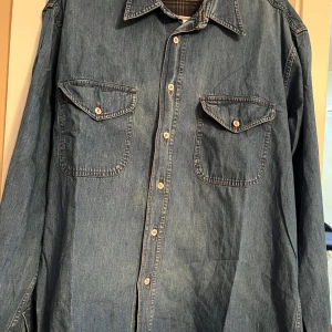 Vintage Blå jeansskjorta från Boss - Snygg blå jeansskjorta från Boss med klassisk krage och knappar framtill. Skjortan har två bröstfickor med knappstängning och ett rutigt foder på insidan. Perfekt för en avslappnad stil.