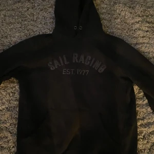 Svart hoodie från Sail Racing - Säljer en svart hoodie från Sail Racing med texten 'SAIL RACING EST. 1977' på framsidan. Den har en klassisk design med huva och dragsko samt en stor magficka. Perfekt för en avslappnad stil.