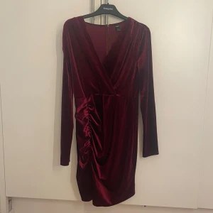 Vinröd sammet klänning - Säljer en elegant vinröd klänning i mjuk sammet. Klänningen har en omlott-design med långa ärmar och en smickrande rynkdetalj på sidan. Perfekt för en stilfull kväll.