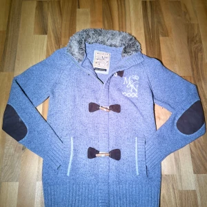 Cardigan med pälskrage / M - Säljer en snygg grå cardigan/jacka med fuskpäls i kragen och broderad logga på bröstet. Finns en liten fläck vid armbågen, storlek M, McNeal, dm mig för fler frågor. Kom gärna med bud