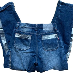 Blå jeans med camouflagedetaljer från FUBU - Snygga baggy blå jeans från FUBU med unika camouflagedetaljer på sidorna och bakfickorna. Byxorna har en loose passform och är perfekta för en avslappnad stil. 38 cm midjemått 100 cm längd 80 cm innebenslängd