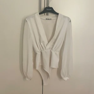Vit omlottbody från boohoo - Snygg vit omlottbody från boohoo med långa ärmar och knytning i midjan. Perfekt för en stilren look. Den har en djup v-ringning och är tillverkad i ett mjukt material. Armarna är i ett mer genomskinligt material.