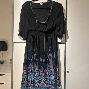 Svart klänning med mönster - Snygg svart klänning med ett färgglatt mönster i blått, lila och grått. Klänningen har trekvartsärmar och en knytning framtill.