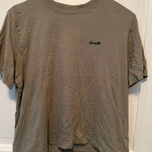 Beige t-shirt från H&M Divided - Säljer en beige t-shirt från H&M Divided med broderad text 'Smile' på bröstet. T-shirten har en rund halsringning och korta ärmar. Perfekt för en avslappnad stil.