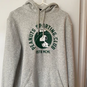 Grå hoodie med Peanuts-tryck - Säljer en grå hoodie med Peanuts Sporting Club-tryck i grönt. Tröjan har en klassisk känguruficka och justerbar huva med snören. Perfekt för en avslappnad stil.