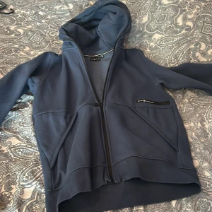 Blå hoodie från Sail Racing - Säljer en snygg blå hoodie från Sail Racing med dragkedja och huva. Den har en stilren design med logotypdetaljer på bröstet och huvan. Perfekt för en casual look. 🌀