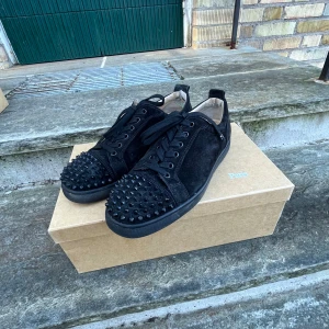 Svarta mockaskor med nitar från Christian Louboutin - Snygga svarta sneakers i mocka från Christian Louboutin med coola nitar på tån och den ikoniska röda sulan. Skorna har snörning och en stilren design som sticker ut.