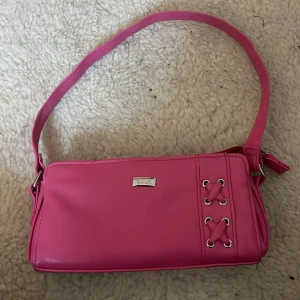 Rosa axelväska med snörning - Snygg rosa axelväska med cool snörningsdetalj på sidan. Perfekt för att ge en färgklick till din outfit. Väskan har en justerbar axelrem och en stilren design med metallogo framtill.