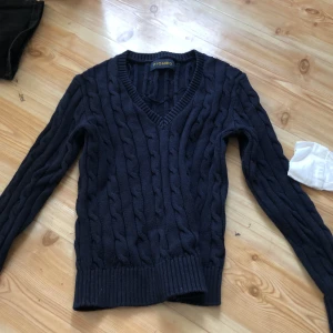 Mörkblå stickad tröja från Piombo - Säljer en snygg mörkblå stickad tröja från Piombo. Tröjan har ett klassiskt flätmönster och en v-ringad hals. Perfekt för kyligare dagar. Passar bra till både jeans och chinos.
