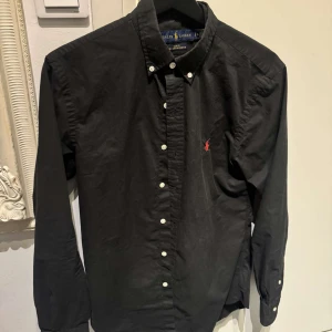 Svart skjorta från Ralph Lauren - Svart skjorta från Ralph Lauren i slim fit. Tillverkad i 100% bomull med stretch. Modell ”Custom Fit Garment Dyed Oxford Shirt Black”, Fint skick.🖤 Skriv om ni vill ha fler bilder. Nypris 1499 kr.