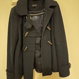 Marinblå kappa från Topshop - Säljer en stilren svart kappa från Topshop med knäppning framtill. Jackan har långa ärmar och en klassisk krage. Perfekt för kyligare dagar och ger en elegant look. Passar bra till både jeans och klänning.