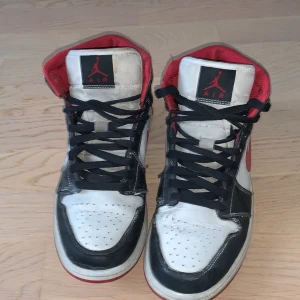 Nike Air Jordans i svart, vitt och rött - Säljer ett par klassiska Nike Air Jordans i svart, vitt och rött. Skorna har snörning och en ikonisk design med hög ankel. Perfekta för sneaker-entusiaster!