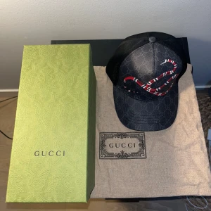 Gucci keps Orm - Snygg svart keps från Gucci med ett iögonfallande rött och vitt ormmotiv. Kepsen har ett diskret mönster med Guccis logotyp och en justerbar baksida för perfekt passform. Perfekt för att ge din outfit en lyxig touch.