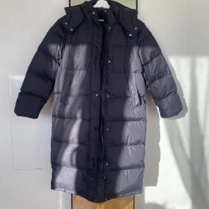 Svart lång vikterjacka - Säljer en svart lång med huva och dragkedja. Jackan har långa ärmar och knappar framtill. Perfekt för kalla dagar.