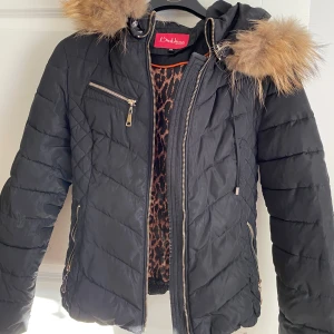 Diabless jacka - Snygg svart quiltad jacka från Diabless med dragkedja och äkta päls på huvan. Jackan har flera praktiska fickor med dragkedjor och ett leopardmönstrat foder som ger en cool touch. Perfekt för kyligare dagar!