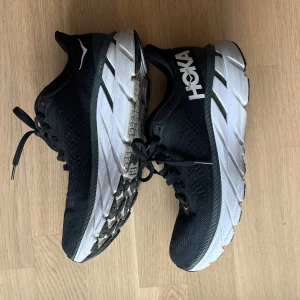 Svarta löparskor från Hoka - Säljer ett par svarta löparskor från Hoka med vit sula och snörning. Skorna har en robust design med märkets logga på sidan och är perfekta för löpning. Modell: clifton 7