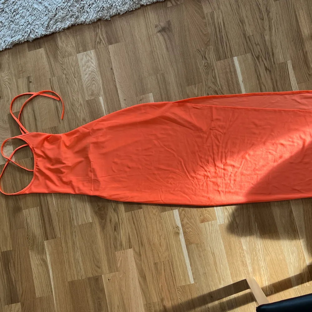 Snygg orange långklänning med smala axelband och öppen rygg. Klänningen har en slits på sidan och snörning i ryggen för en justerbar passform. Perfekt för sommarens varma dagar.. Mekot.