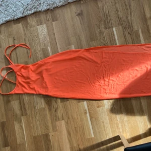 Orange långklänning med snörning - Snygg orange långklänning med smala axelband och öppen rygg. Klänningen har en slits på sidan och snörning i ryggen för en justerbar passform. Perfekt för sommarens varma dagar.