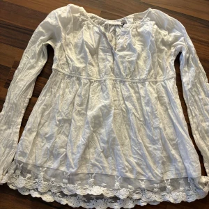 Vit blus från Lagar 157 - Säljer en söt vit blus från Lager 157 med långa ärmar och fina spetsdetaljer vid nederkanten. Blusen har en v-ringad design med knappar framtill. Perfekt för en romantisk look!