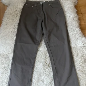 GråBruna byxor från Cappuccini nya - Snygga bruna byxor från Cappuccini med broderad detalj på bakfickan. Byxorna har en klassisk design med knappar och dragkedja framtill. Perfekta för en stilren look.
